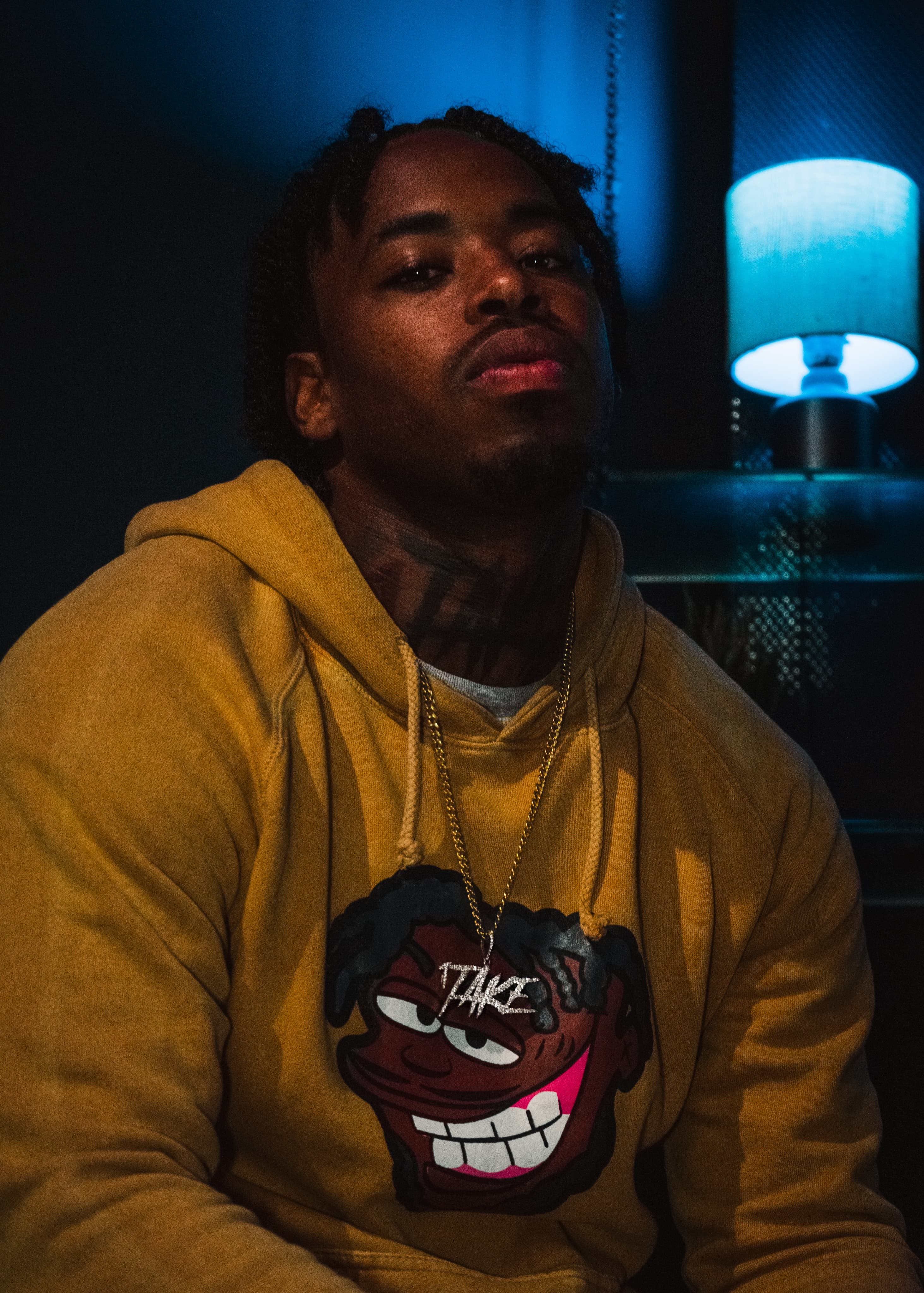 1TakeQuan in a mustard hoodie with the character logo, blue lamp behind